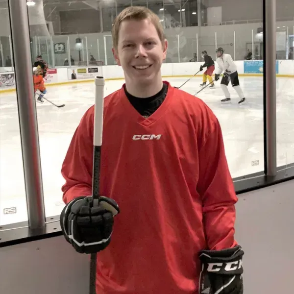 Man in red hockey jersey smiles holding a hockey stick and glove on the ice rink.
