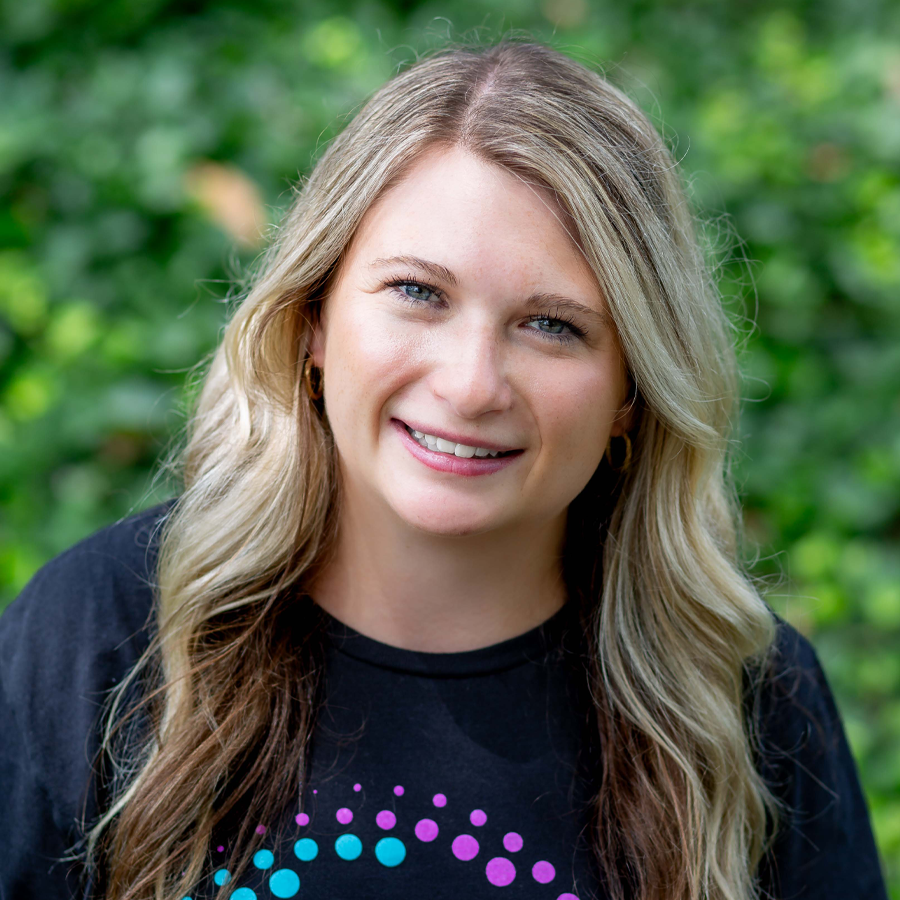 Woman with blonde hair smiles, wearing a black shirt with a colorful design, outdoors with greenery.
