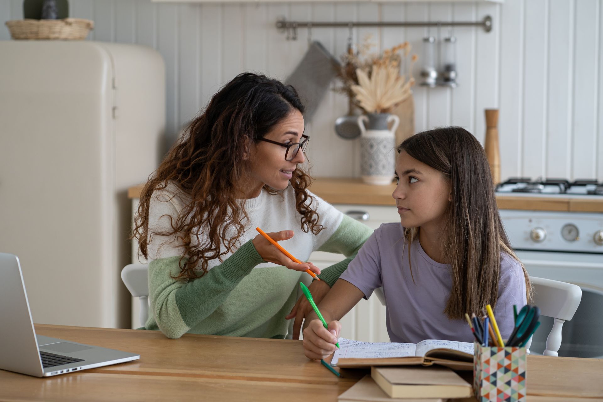 Woman helping a child with schoolwork at a kitchen table. They are both looking at a notebook.