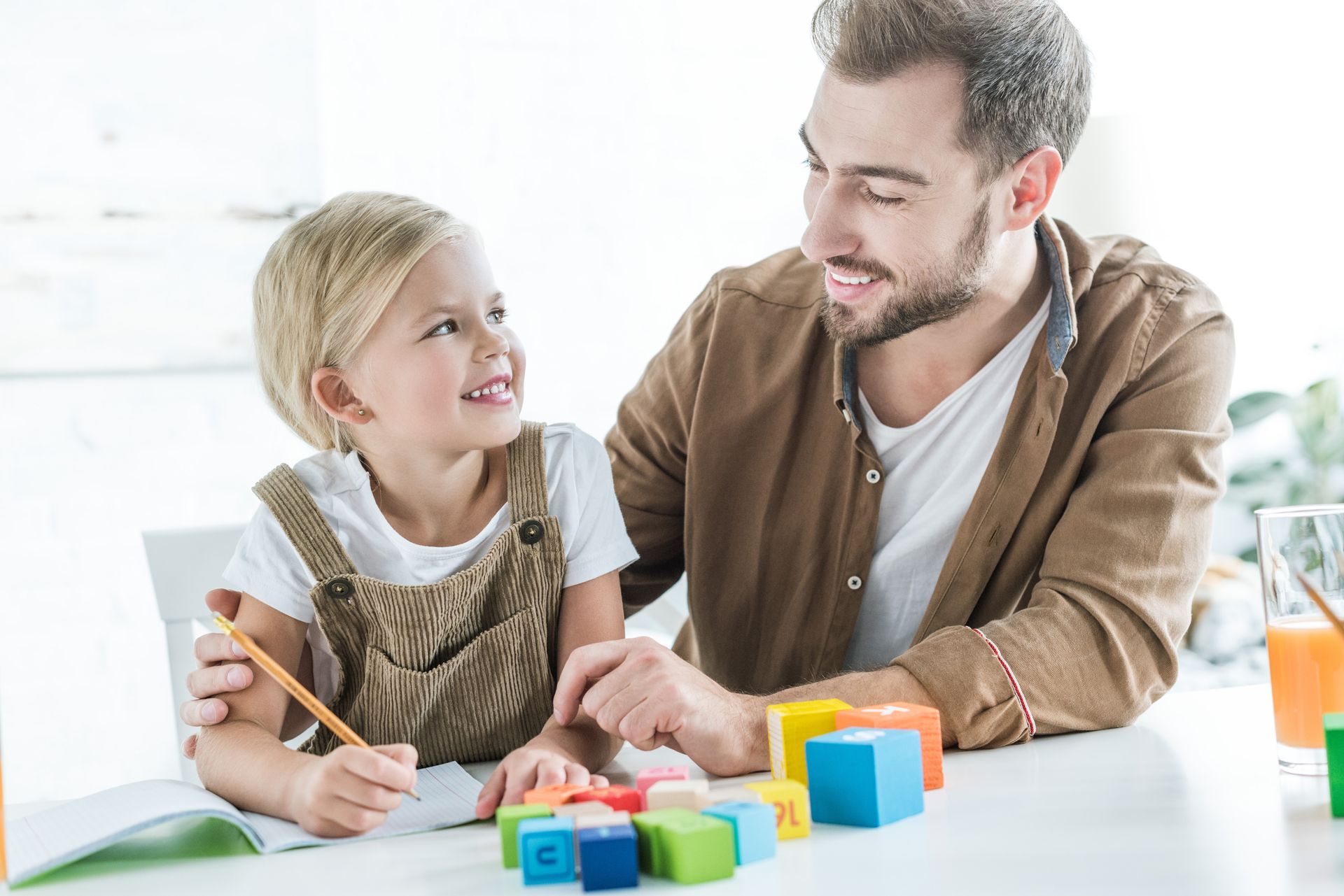 A man and a child looking at each other at a table with toys and a drawing book.