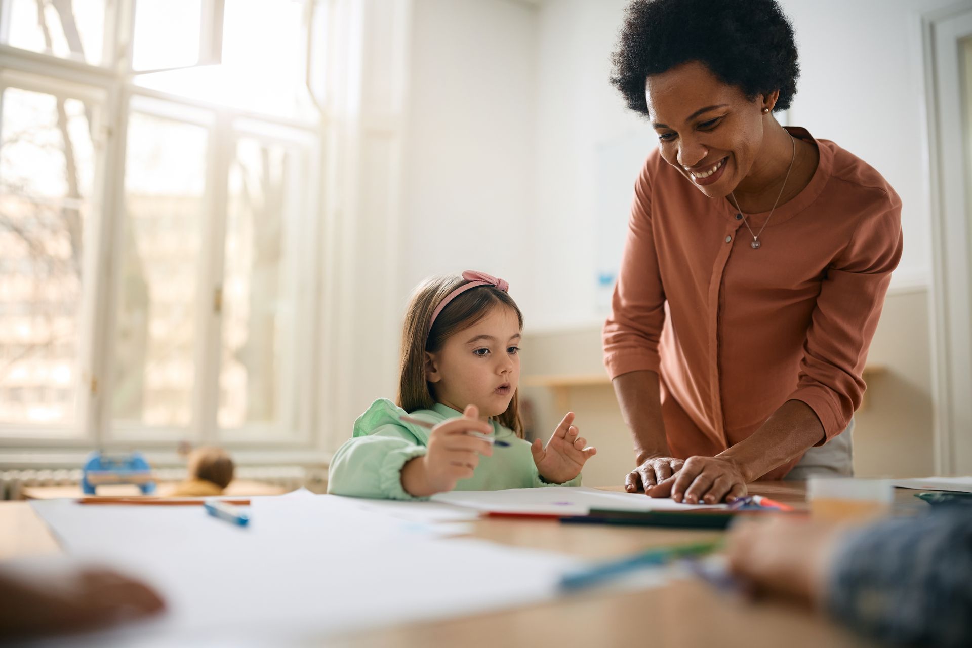 Woman helps a child with drawing at a table, sunny room with a window in the background.