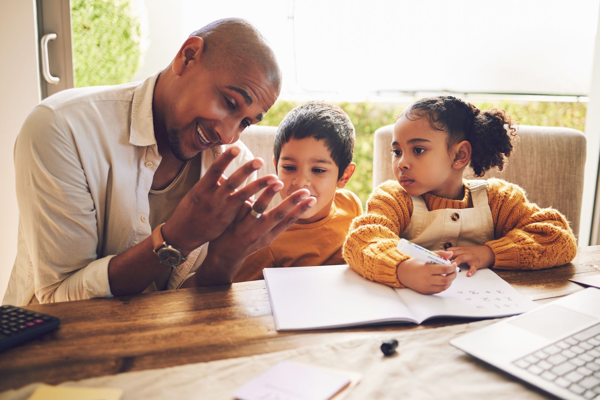 Man helps two children with schoolwork at a table. They look at a notebook, smiling.