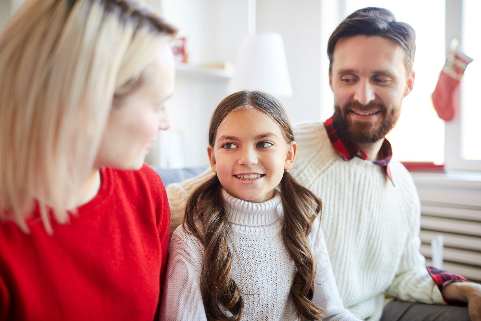 Family of three, daughter in center, parents on either side, indoors. Smiling and looking at each other, Christmas stocking in background.