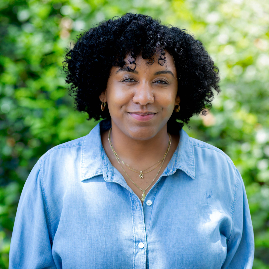 Woman with curly dark hair, smiling, wearing a cream-colored collared shirt, against a white wall.
