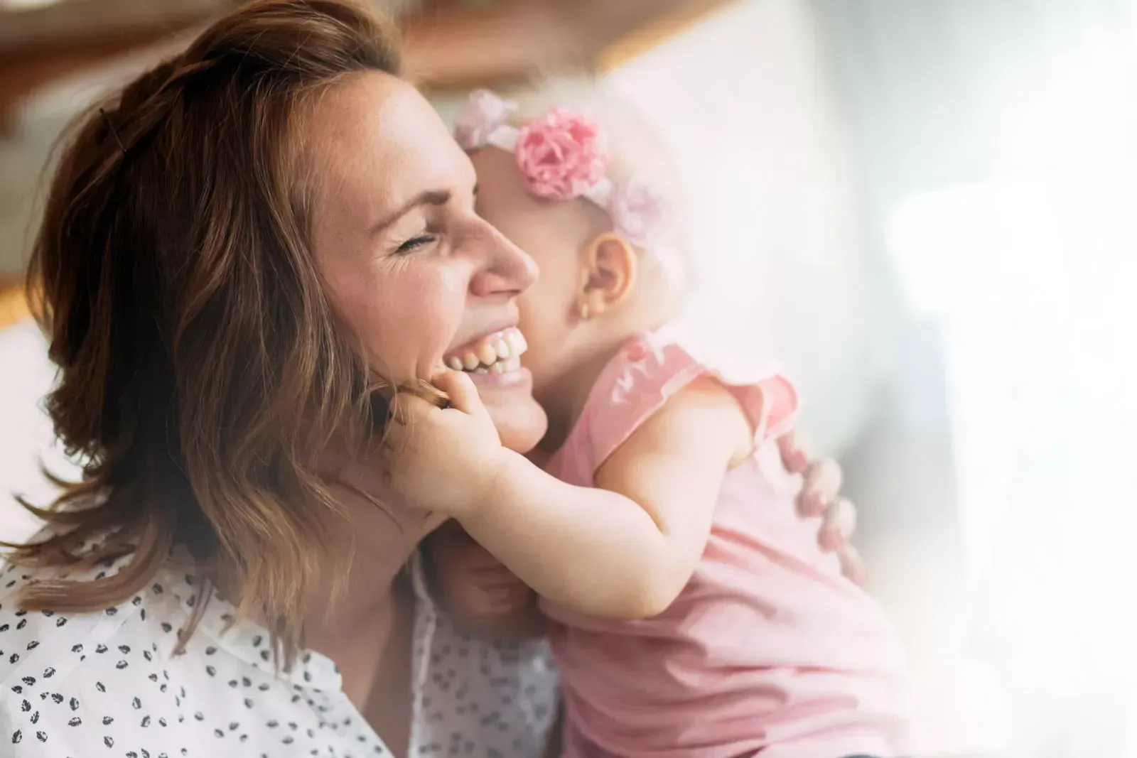 Smiling woman and baby, playfully touching faces. Baby wears pink and floral headband.