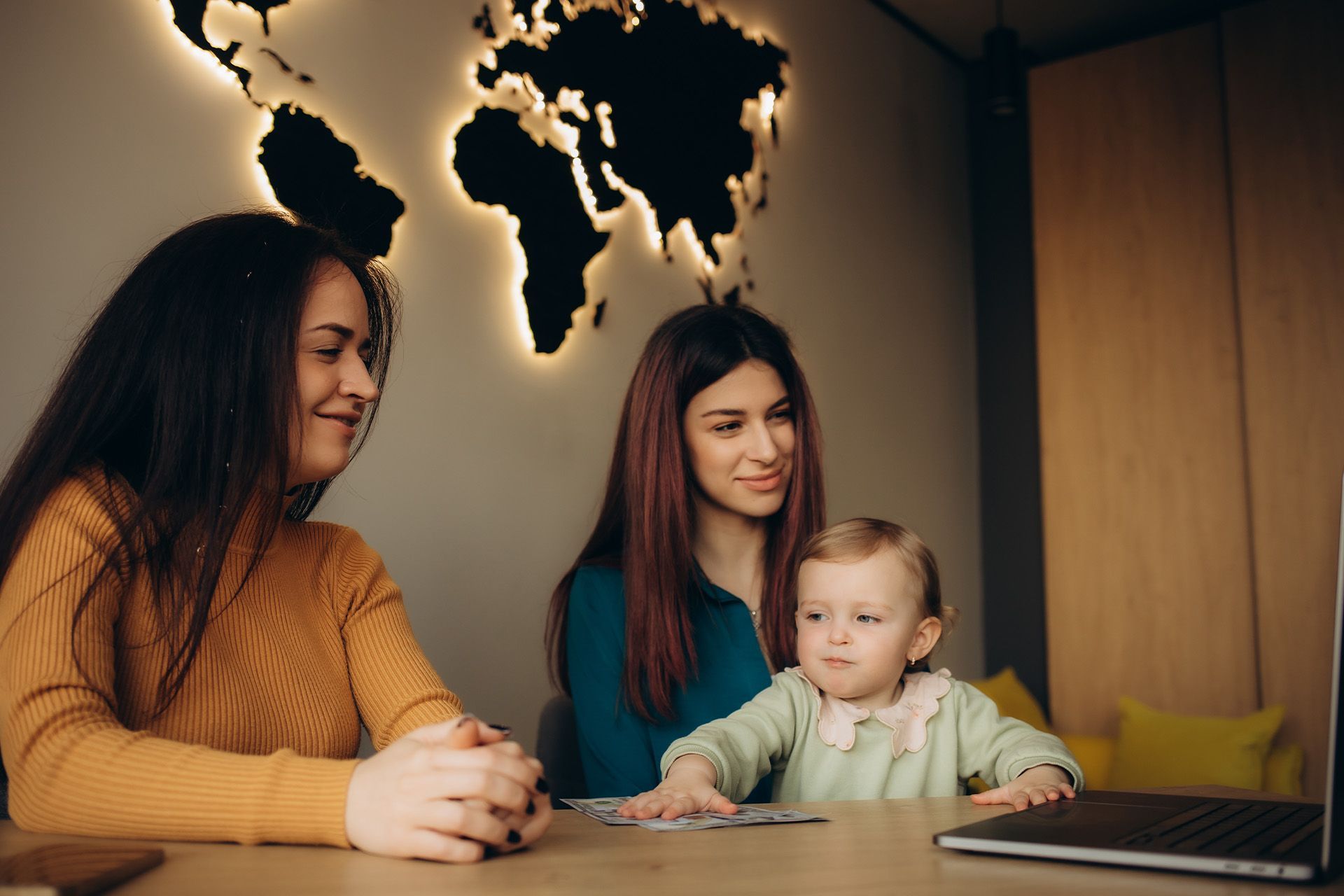 Two women and a baby looking at a laptop. They sit at a table in front of a world map.