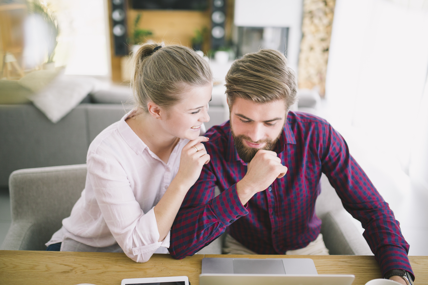 Woman leans on man's shoulder, looking at laptop. Both are smiling in a home office setting.