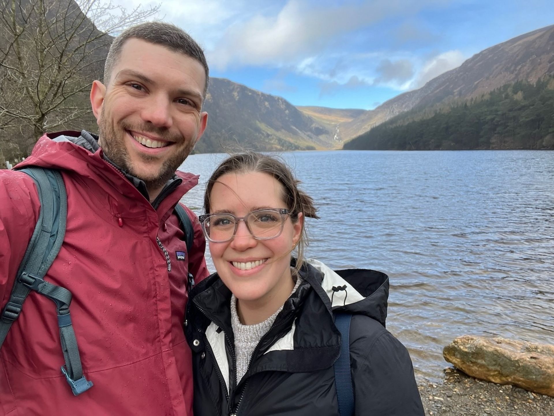 Couple smiles for a selfie at a lake with mountains in the background.