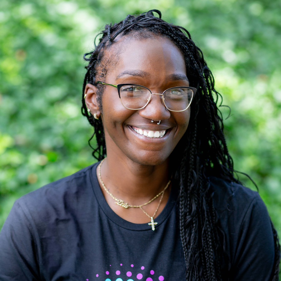 Smiling Black woman with braids, glasses, and nose ring, in front of green foliage.