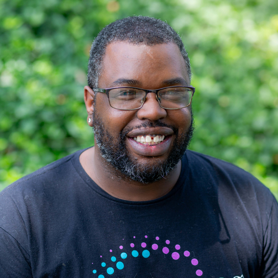 Smiling Black man with glasses, black beard, and black t-shirt outdoors in front of leafy green background.