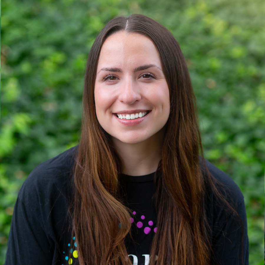 Woman with long brown hair smiling at the camera, wearing a black shirt, in front of green foliage.