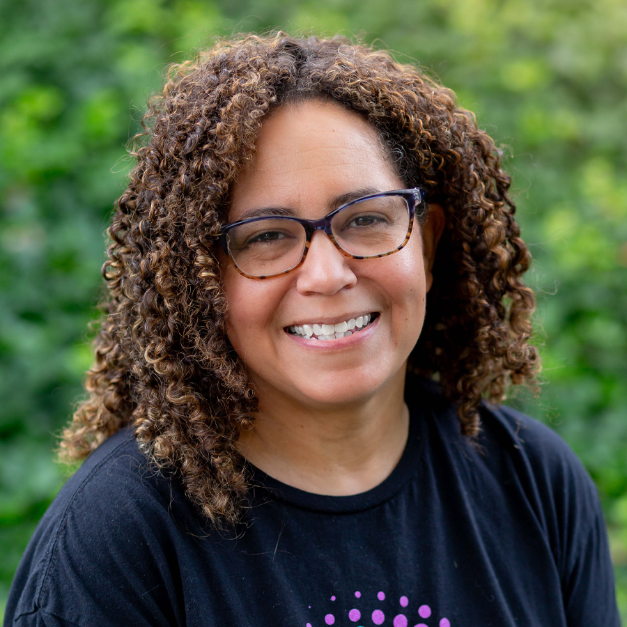 Woman with curly brown hair and glasses smiles outdoors.