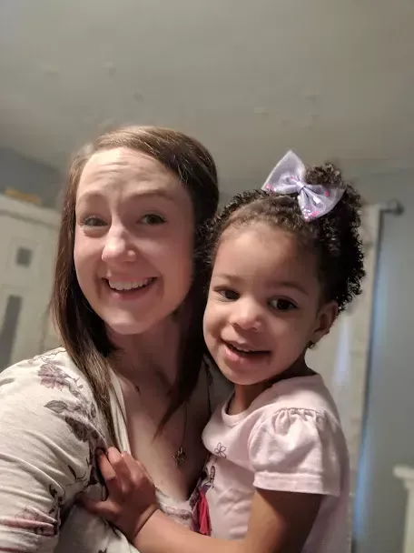 Woman smiles, holds small girl with a purple bow; both smile at the camera indoors.