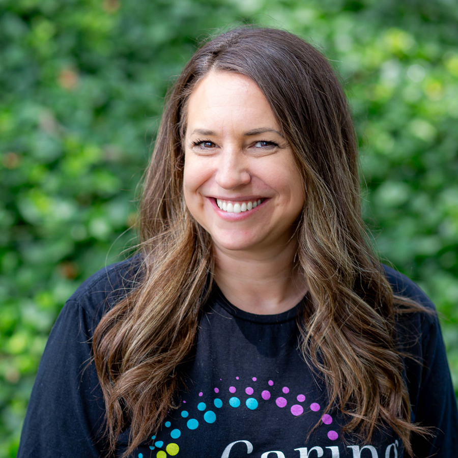 Woman with long brown hair smiles, wearing a black shirt with colorful dots, outdoors with green foliage.