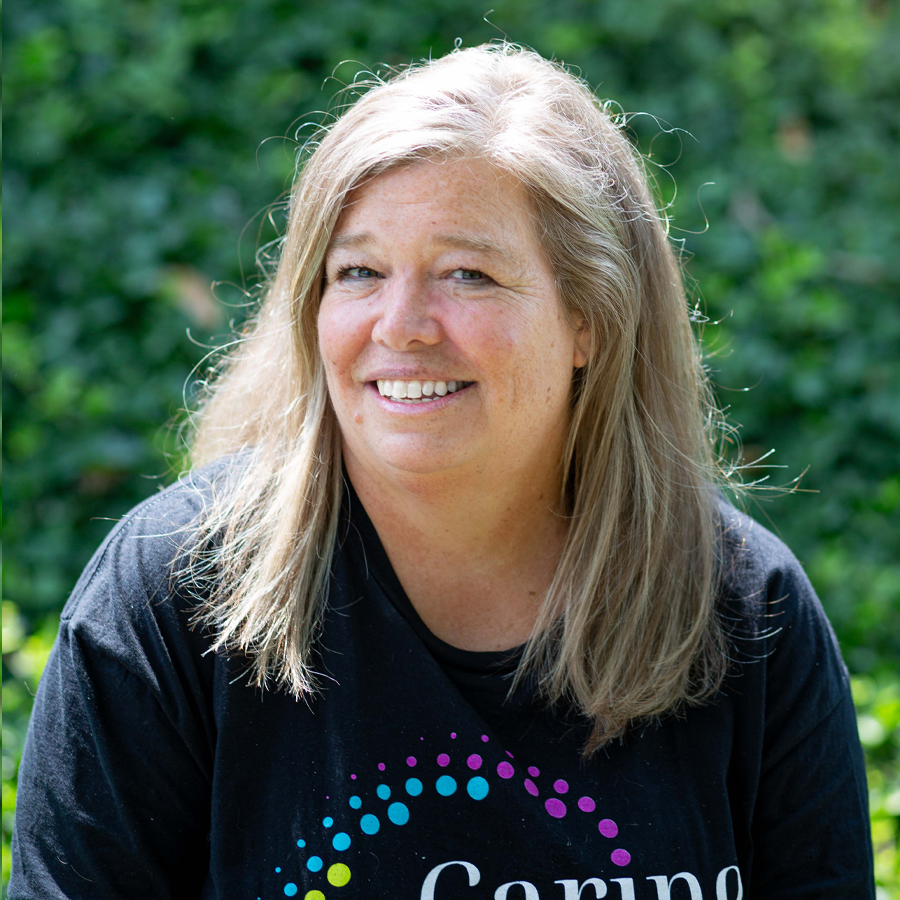 Woman with blonde hair wearing a black t-shirt smiles in front of green foliage.