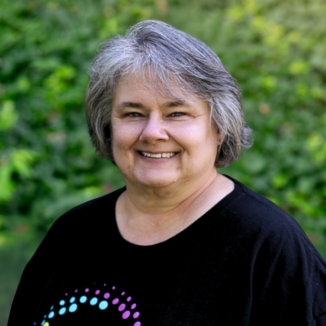 Woman with glasses, smiling, wearing a black and patterned top, in front of a light gray background.