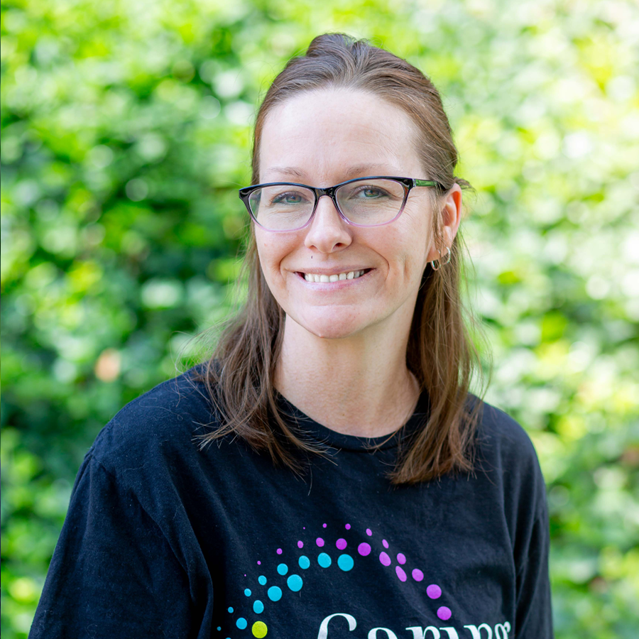 Woman with glasses smiles outdoors, wearing a black shirt with colorful design.