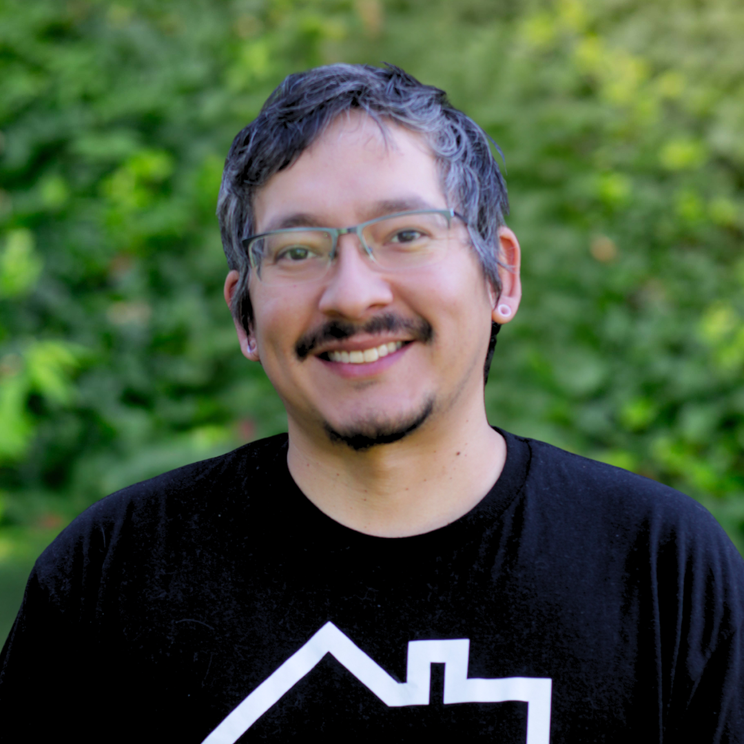 Person with glasses smiles, wearing green sweater, standing in front of a white wall.