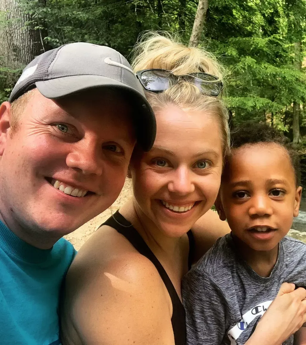 Family smiling together outdoors: man in hat, woman with blonde bun, young Black child.