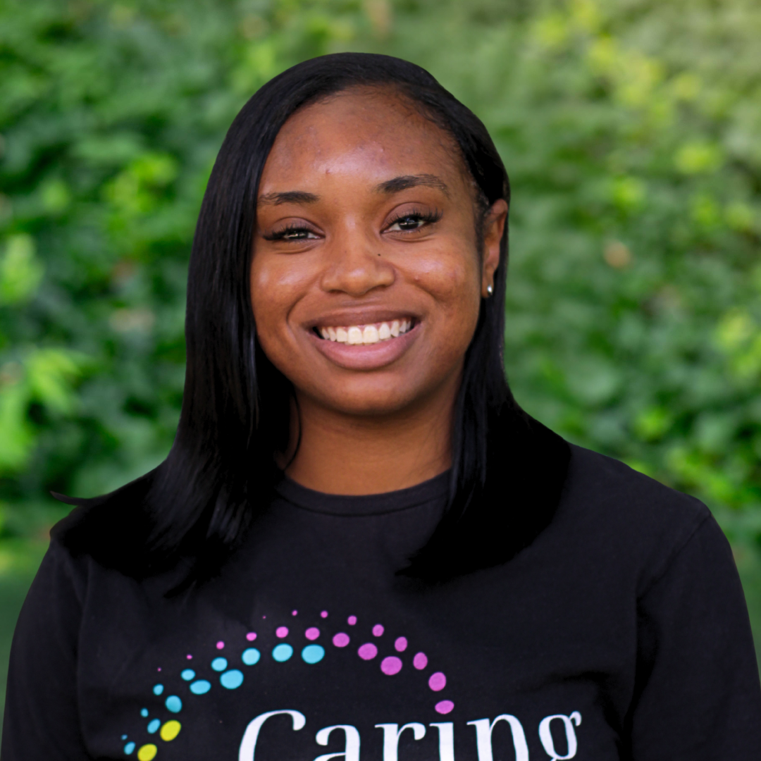 A young Black woman smiles at the camera, wearing a gray sweater and a floral tee.