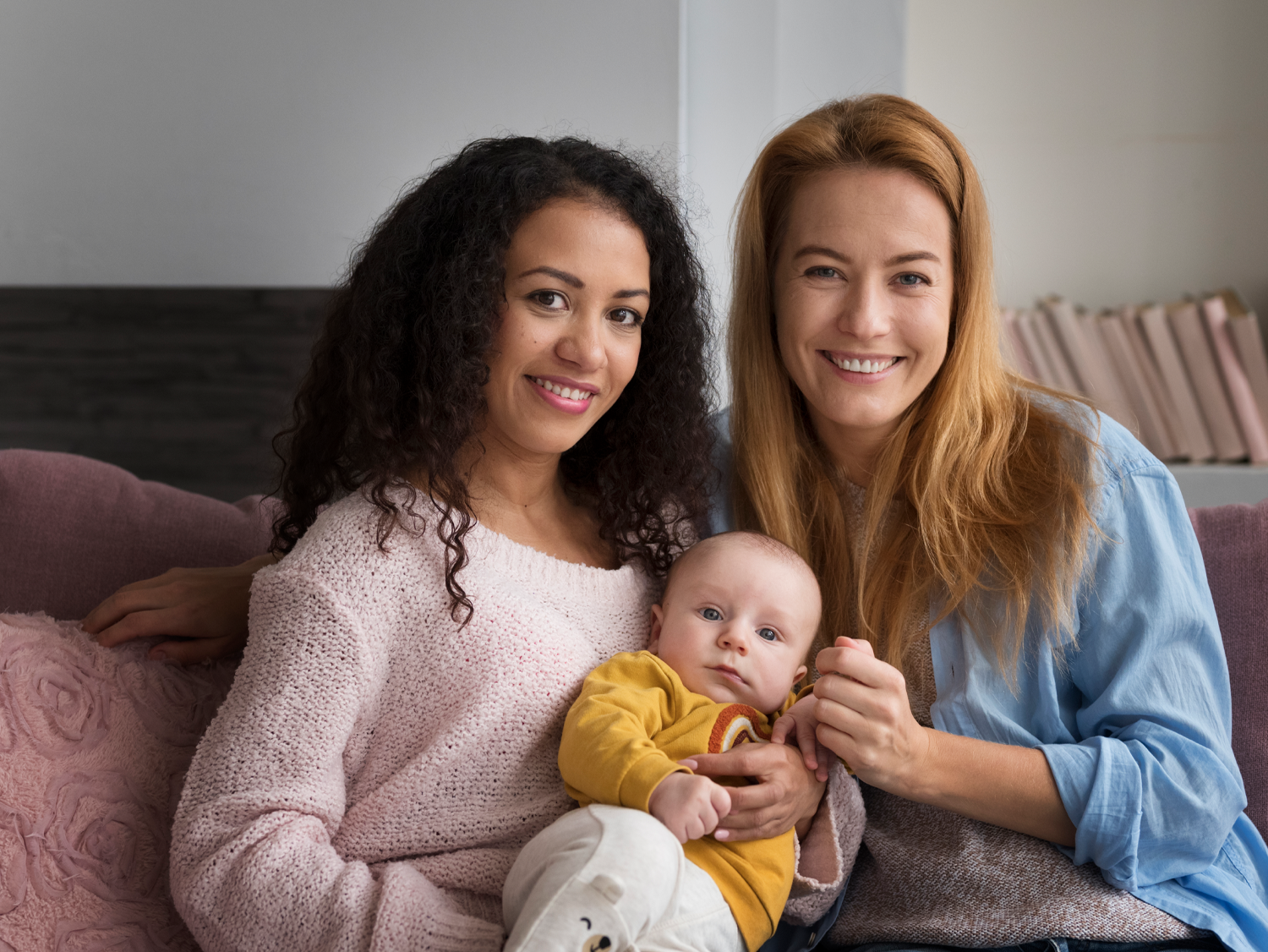 Two women of diverse ethnicities and baby smiling on a couch.