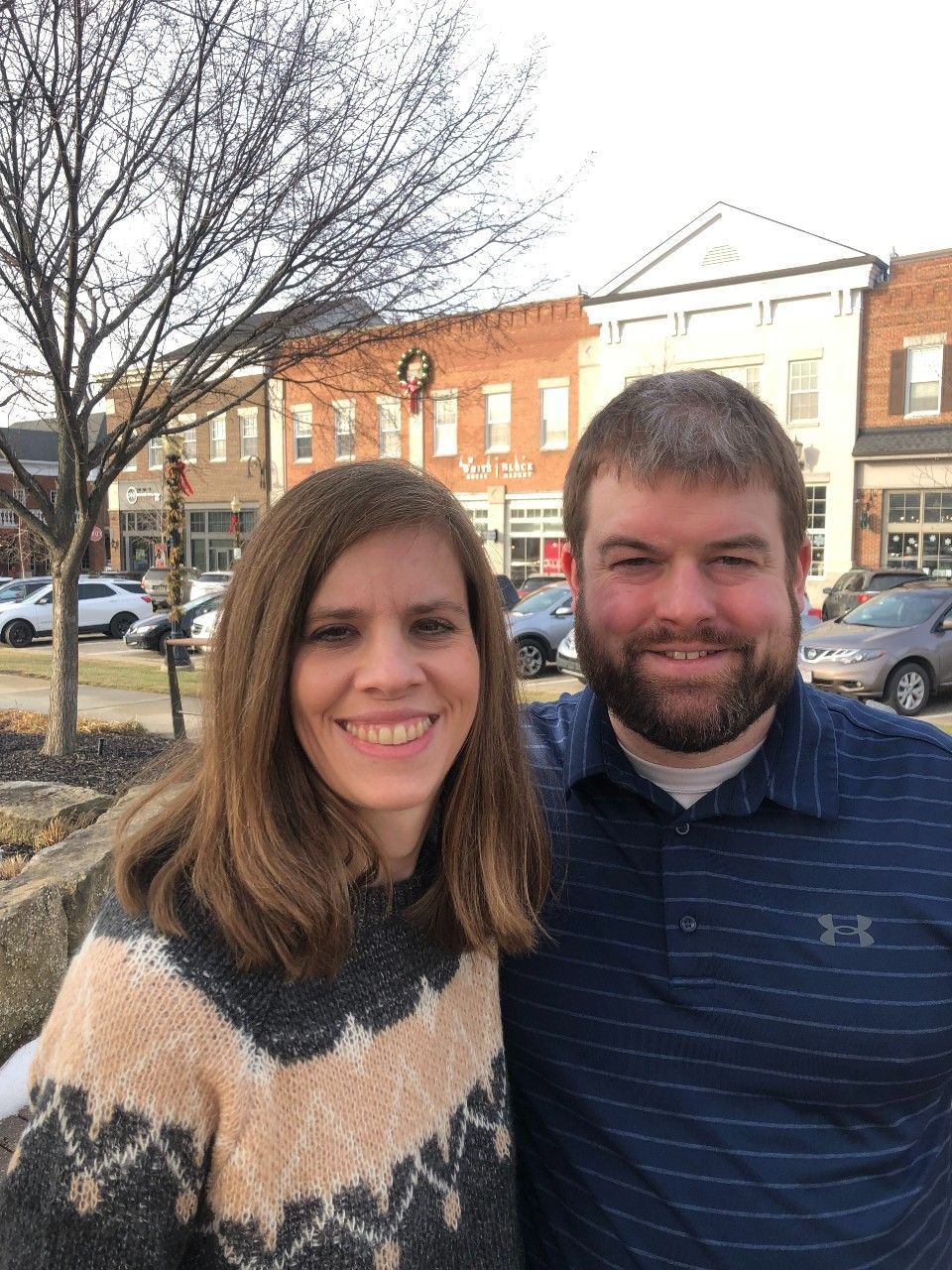 Woman and man smiling outdoors in front of brick buildings. Woman wears a patterned sweater, man has a beard.
