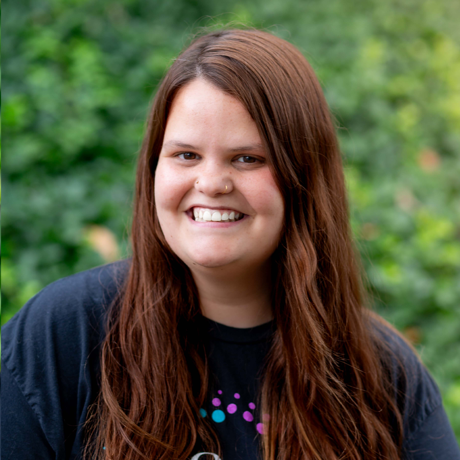 Woman with long brown hair smiles broadly at the camera outdoors, wearing a black shirt.