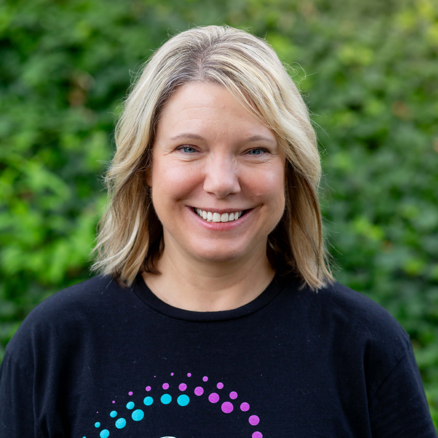 Blonde woman smiling, wearing a black shirt with a colorful design, outdoors in front of green foliage.