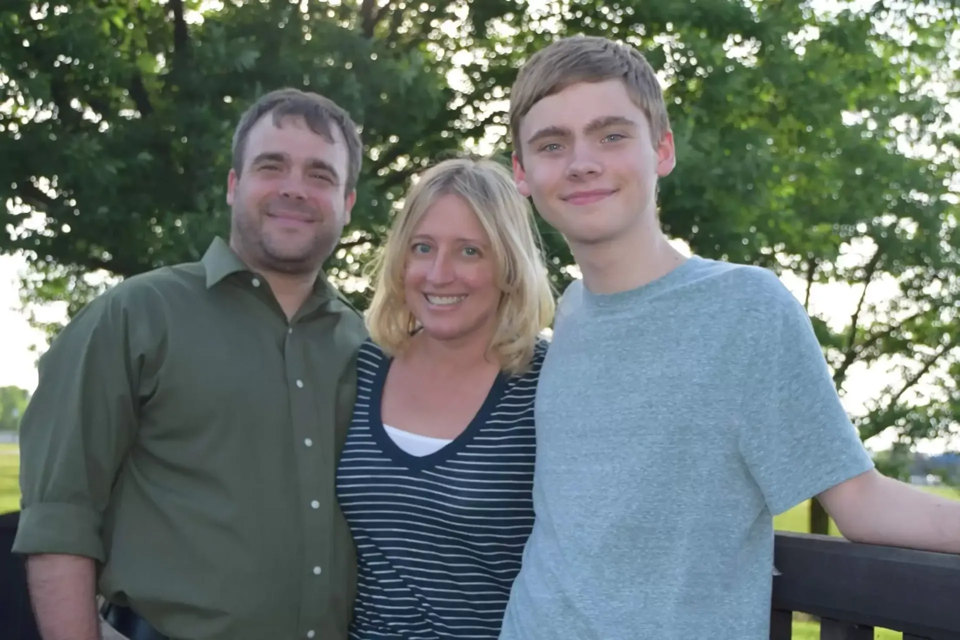 Family of three smiling outdoors; man in green shirt, woman in striped top, teen in blue shirt.