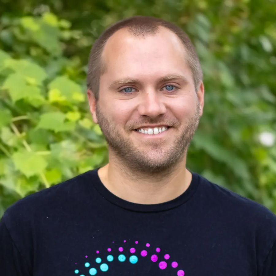 Man with glasses and grey hoodie smiles in front of a white wall.