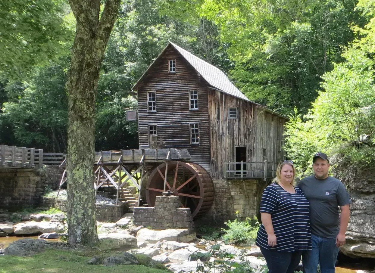 Couple poses in front of an old wooden grist mill with a waterwheel in a wooded area.