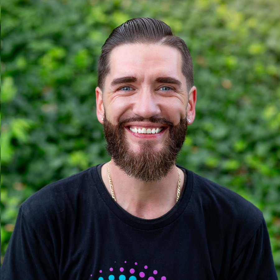 Smiling man with a beard in front of green foliage. He has a dark t-shirt and a gold chain.