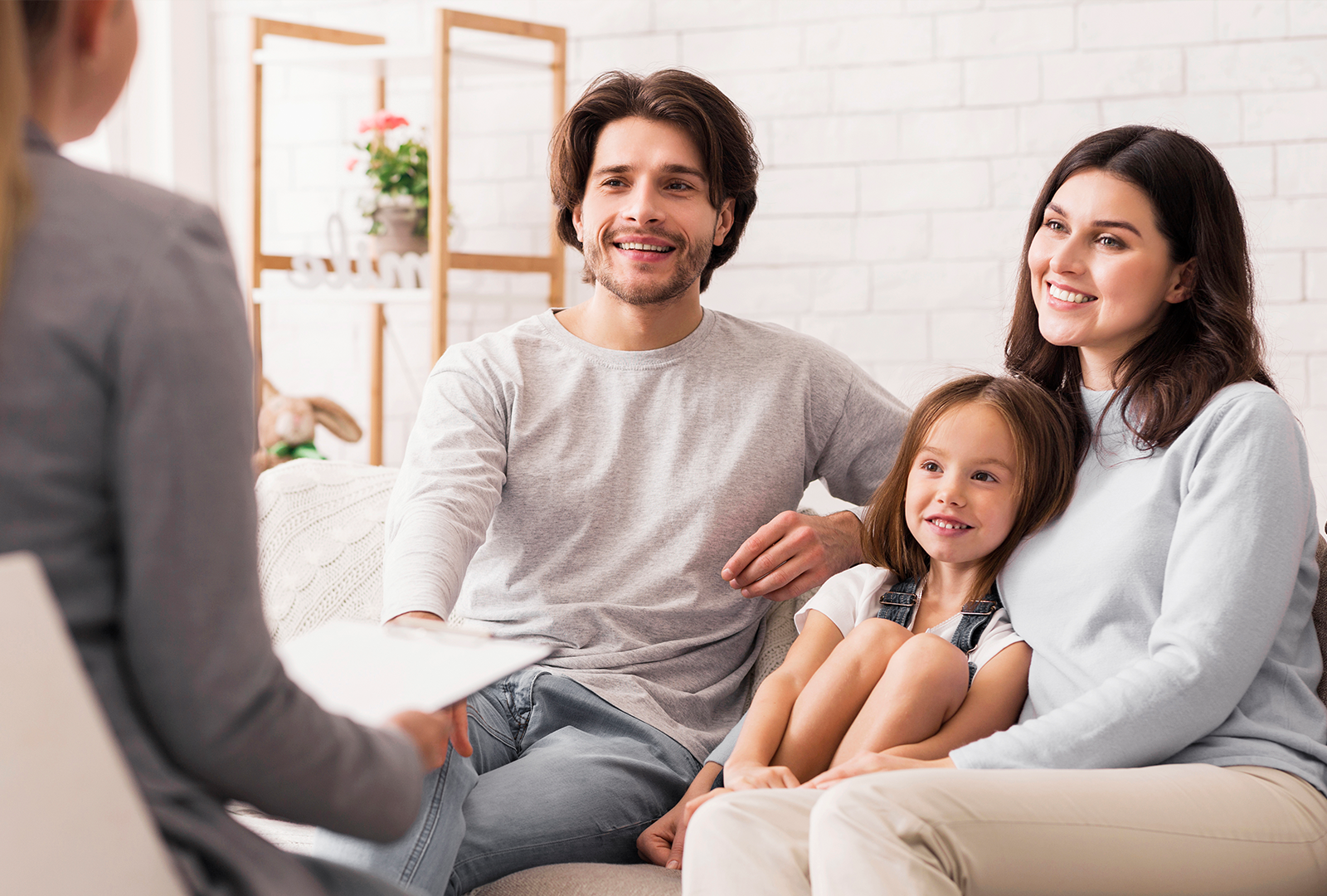 Family of three smiling, sitting on a couch, talking to a woman. White brick wall background.