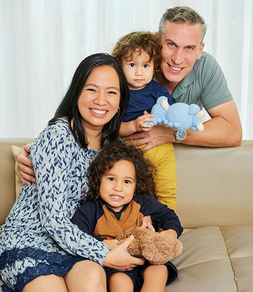 Family of four, smiling together indoors. Mother seated, father behind holding child. Another child in front.