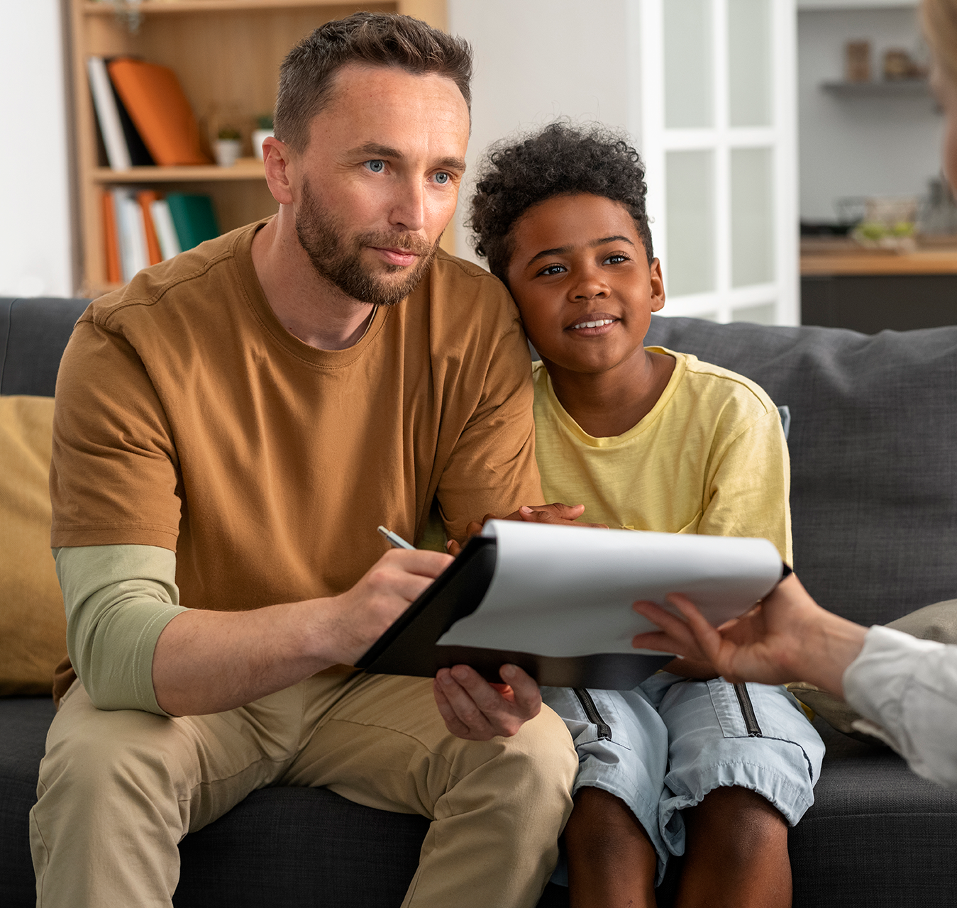 Father and child on a sofa looking at paperwork with a person; living room setting.