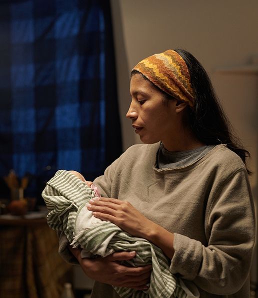 Woman holding a swaddled baby. She wears a headband and looks down. Soft lighting.