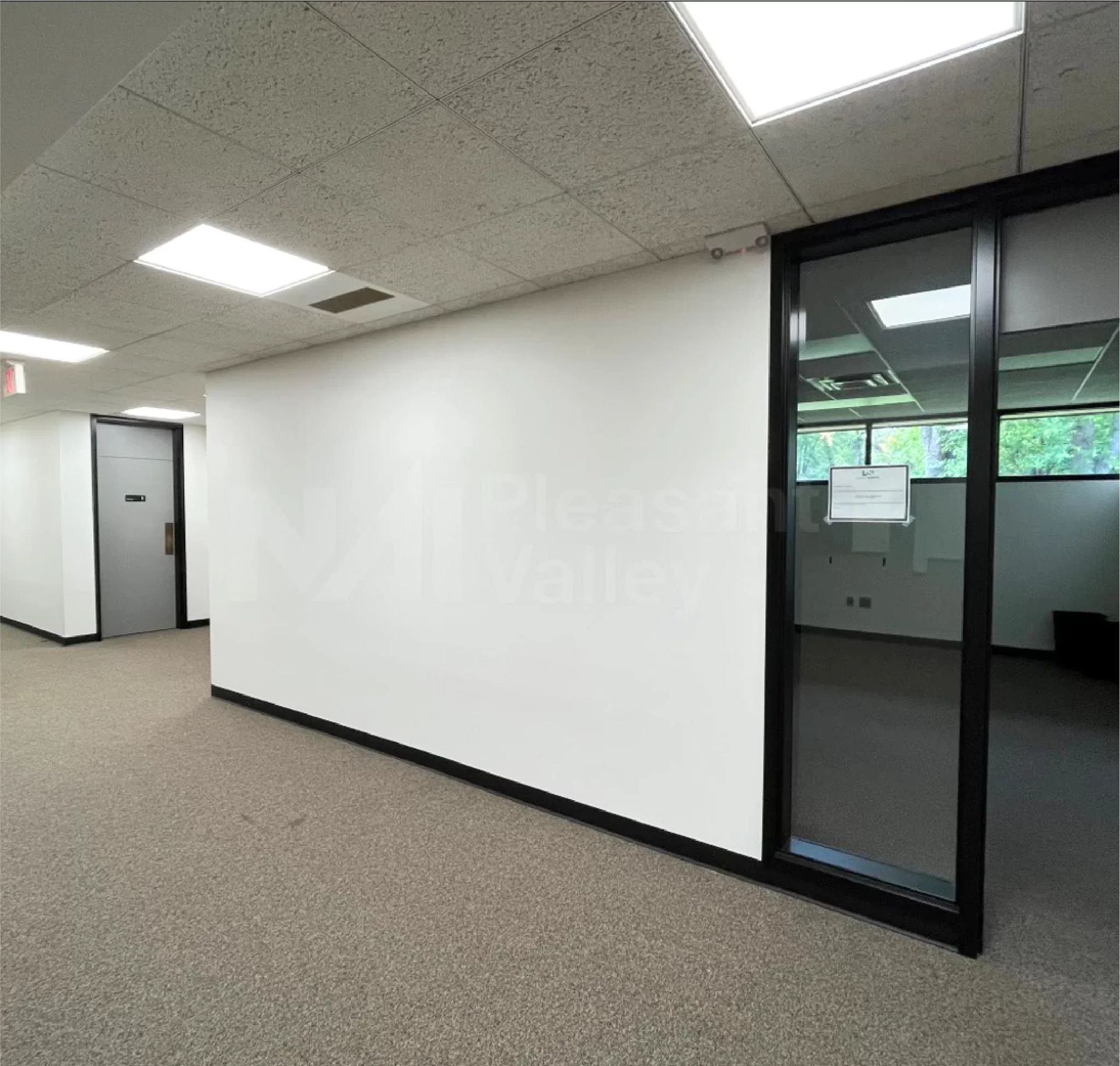 Empty office hallway with white walls, carpeted floor, and glass door to another room.