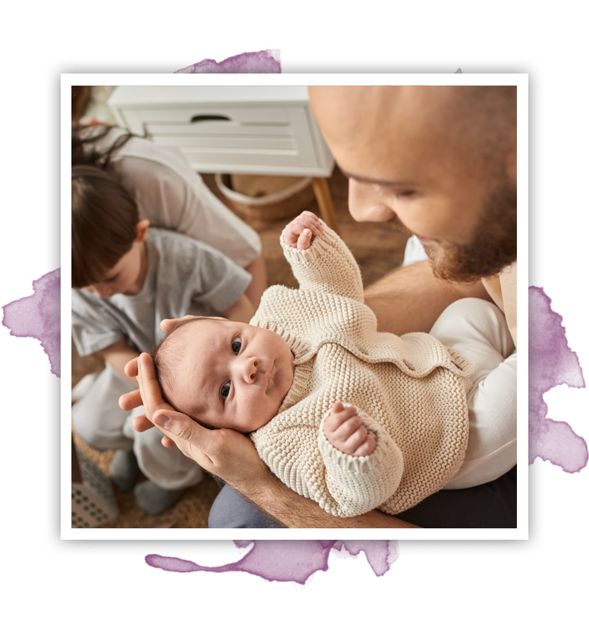 Man holds newborn, looking up. Older sibling and parent in background. Neutral tones, indoor setting.