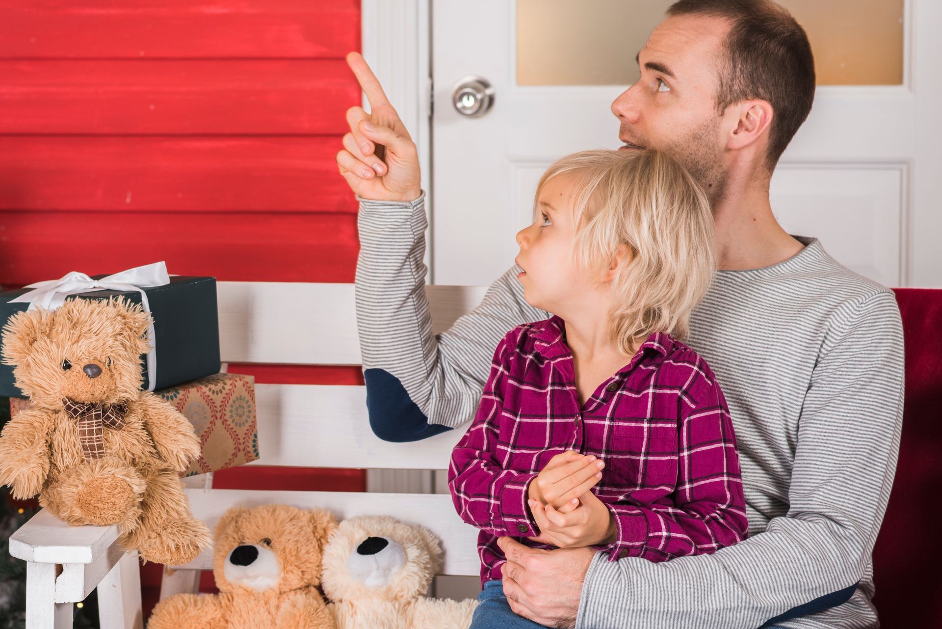 Father points upward, holding a young girl. They sit on a bench with teddy bears, red wall in background.