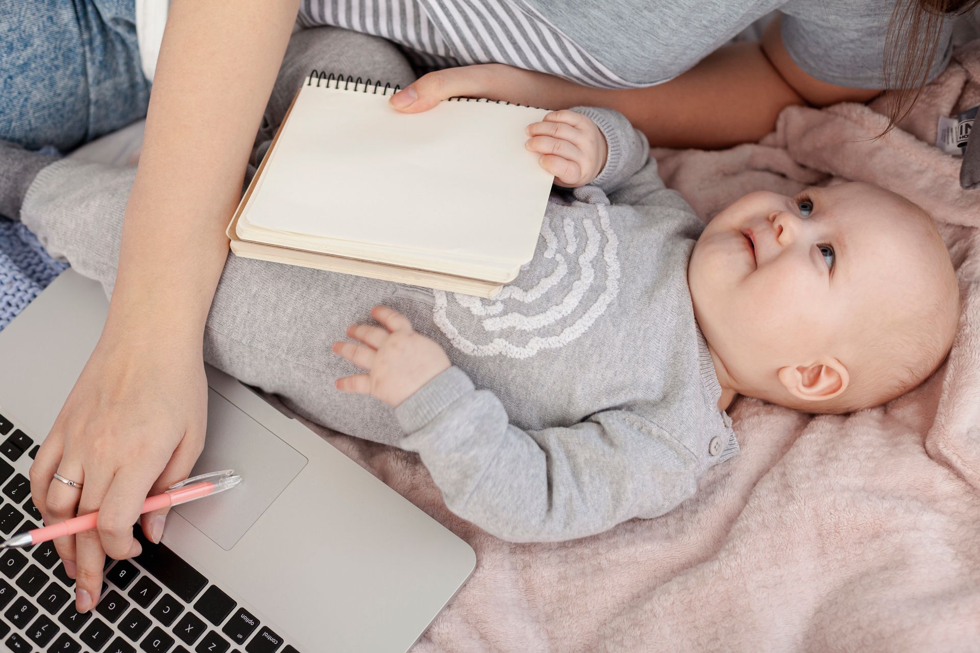 Woman working on laptop, holding baby, writing in notebook on bed.