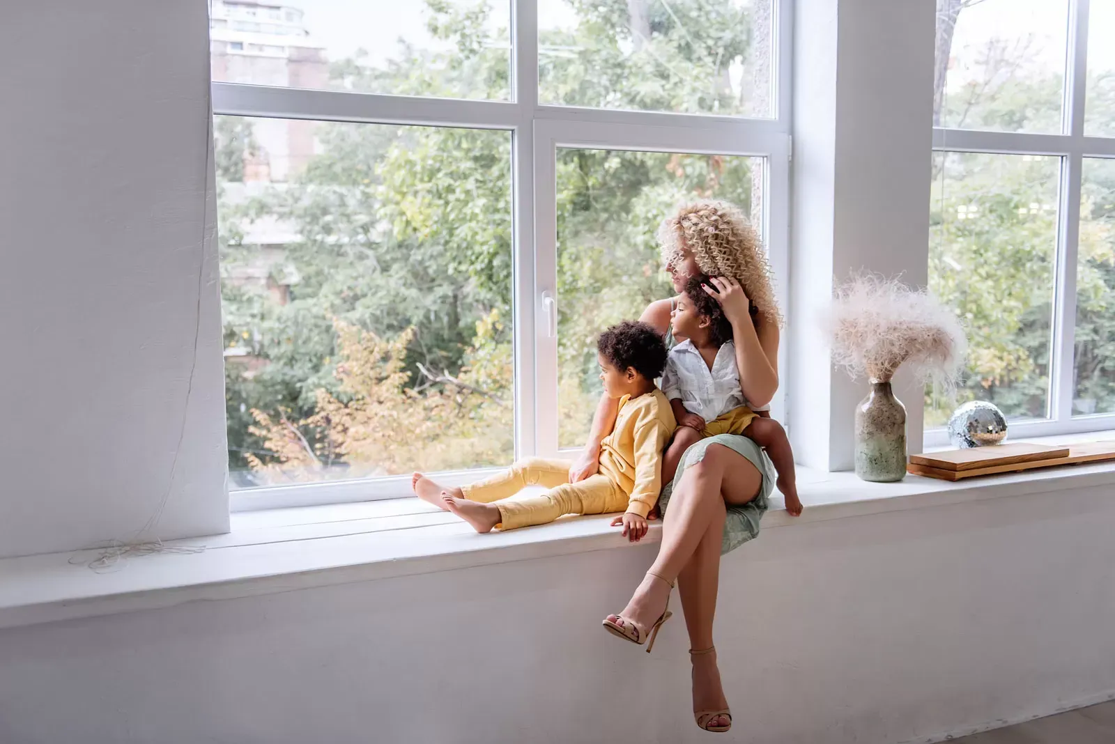 Woman with curly hair sits on a windowsill with two children, looking out. White walls, large window, trees.