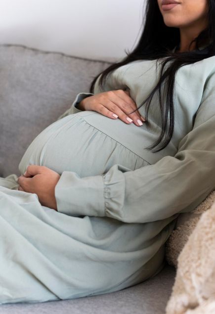 Pregnant woman in sage green dress gently holding her baby bump while sitting on a couch.
