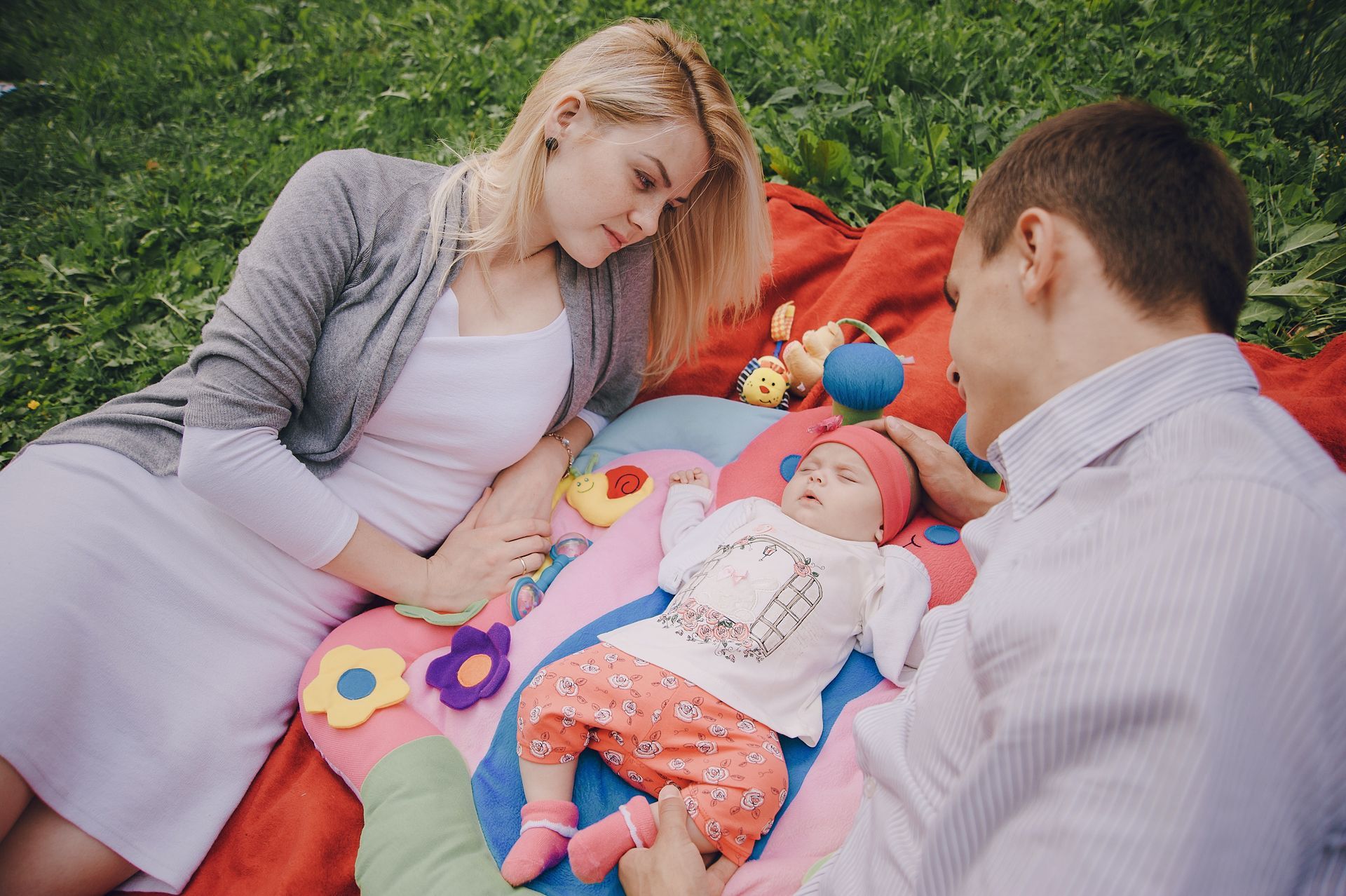 Parents and baby on a picnic blanket in the grass. The baby wears a hat and pink pants, parents are looking at the baby.
