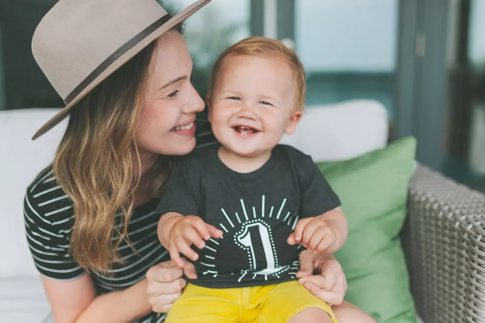 Woman with hat smiles at a toddler on a couch. The boy laughs wearing a shirt with 