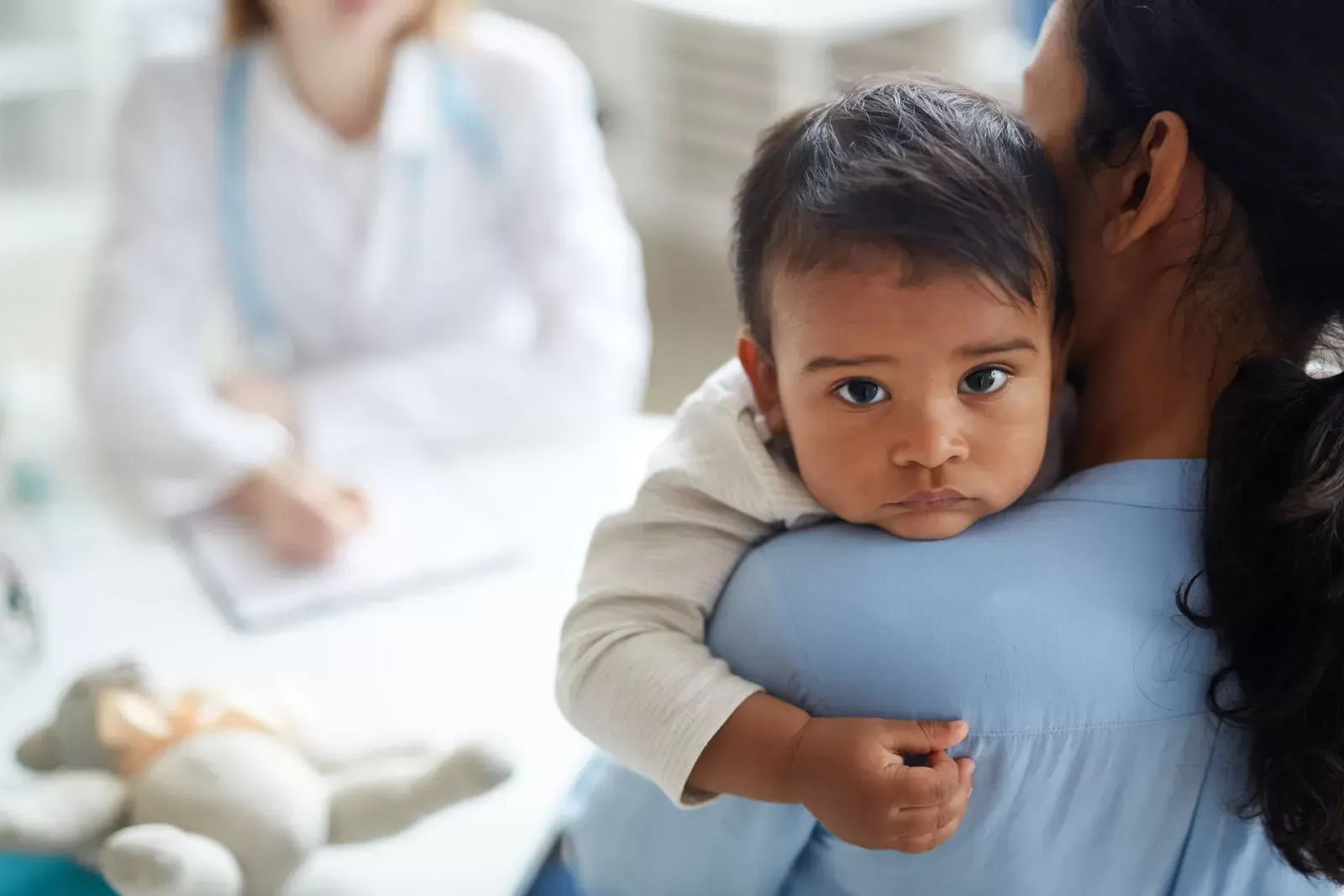 A baby looks sad while being held by a parent, with a doctor in the background.