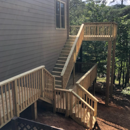 Wooden deck and stairs on the side of a gray house, set in a wooded area with trees.