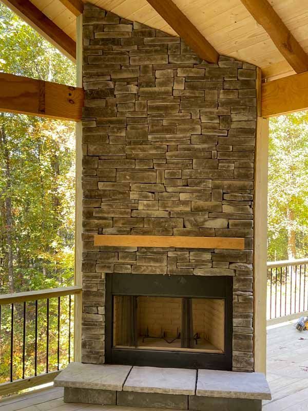 A stone fireplace sits on a wooden porch, featuring a wood mantel and a light-colored stone hearth.