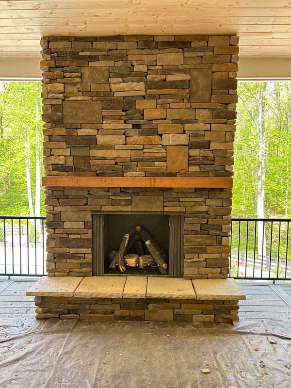 Stone fireplace with wood mantel, built on a patio with a metal railing, overlooking trees.