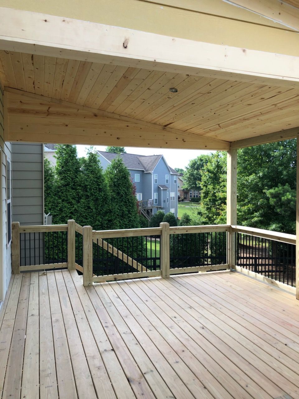 Covered wooden deck with black railings and a view of trees and houses.
