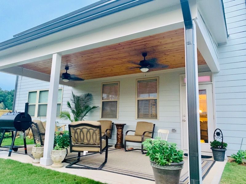 A covered patio with a wooden ceiling, two ceiling fans, outdoor seating, a grill, and potted plants next to a house.